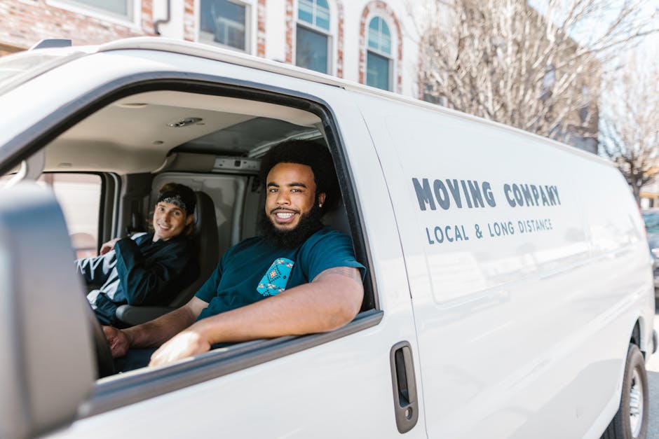 Two movers, dressed in dark clothing with protective eyewear, are carefully loading a large piece of furniture covered in dark green fabric into the back of a white cargo van on a paved street. The va