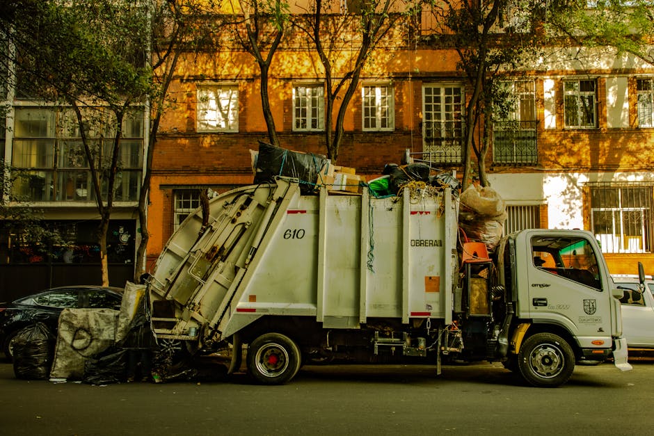 A black and white photograph of a waste disposal truck parked outside a residential building in an urban setting. The truck is positioned alongside the pavement, with the cab on the right and the larg