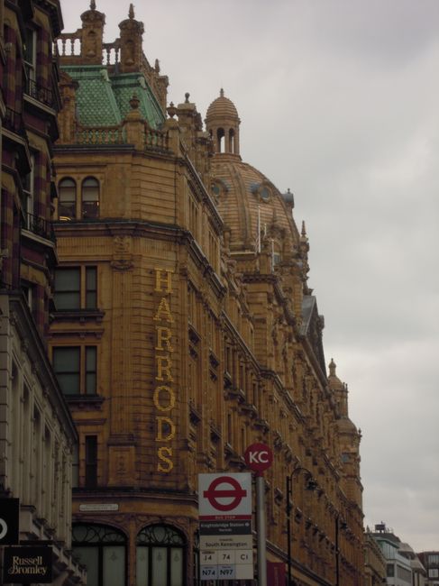 A large historic building with ornate architectural details, including decorative stonework, domed towers, and intricate roof structures, situated on a city street under an overcast sky. The building'