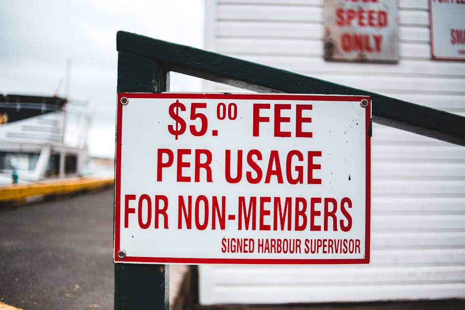 Close-up view of a rectangular white sign with red text displaying a fee of $5.00 per usage for non-members, mounted with four metal bolts on a green wooden post at a harbour or marina. In the backgro